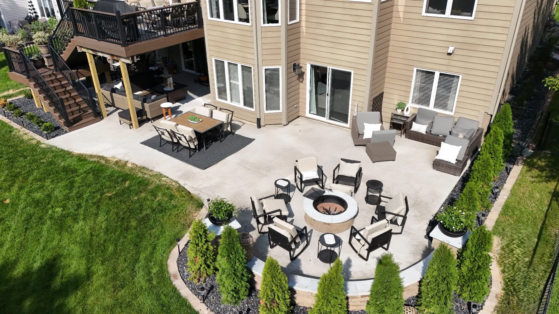 Aerial view of an outdoor patio featuring a seating area around a fire pit, dining space, and landscaped surroundings with green grass and decorative plants.