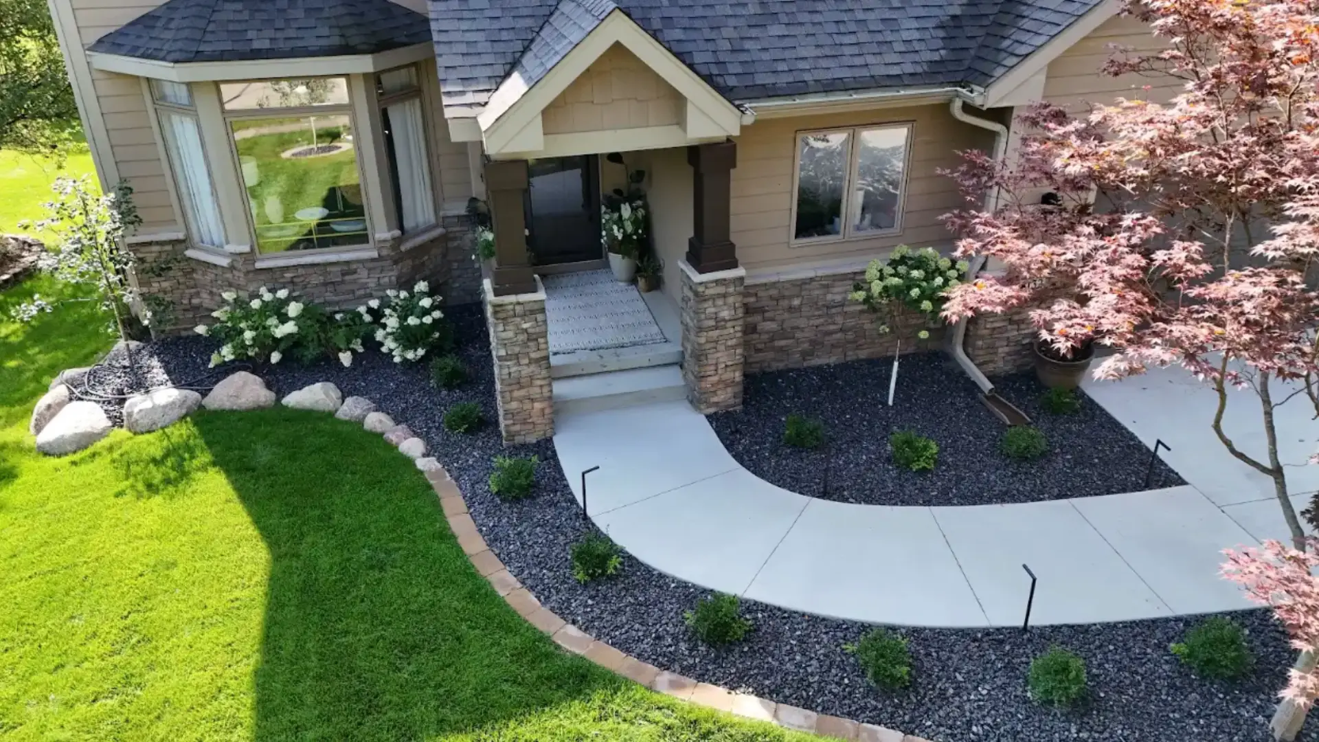 Aerial view of a modern home's entrance featuring a landscaped front yard with green grass, flower beds, and a stone pathway leading to the porch.