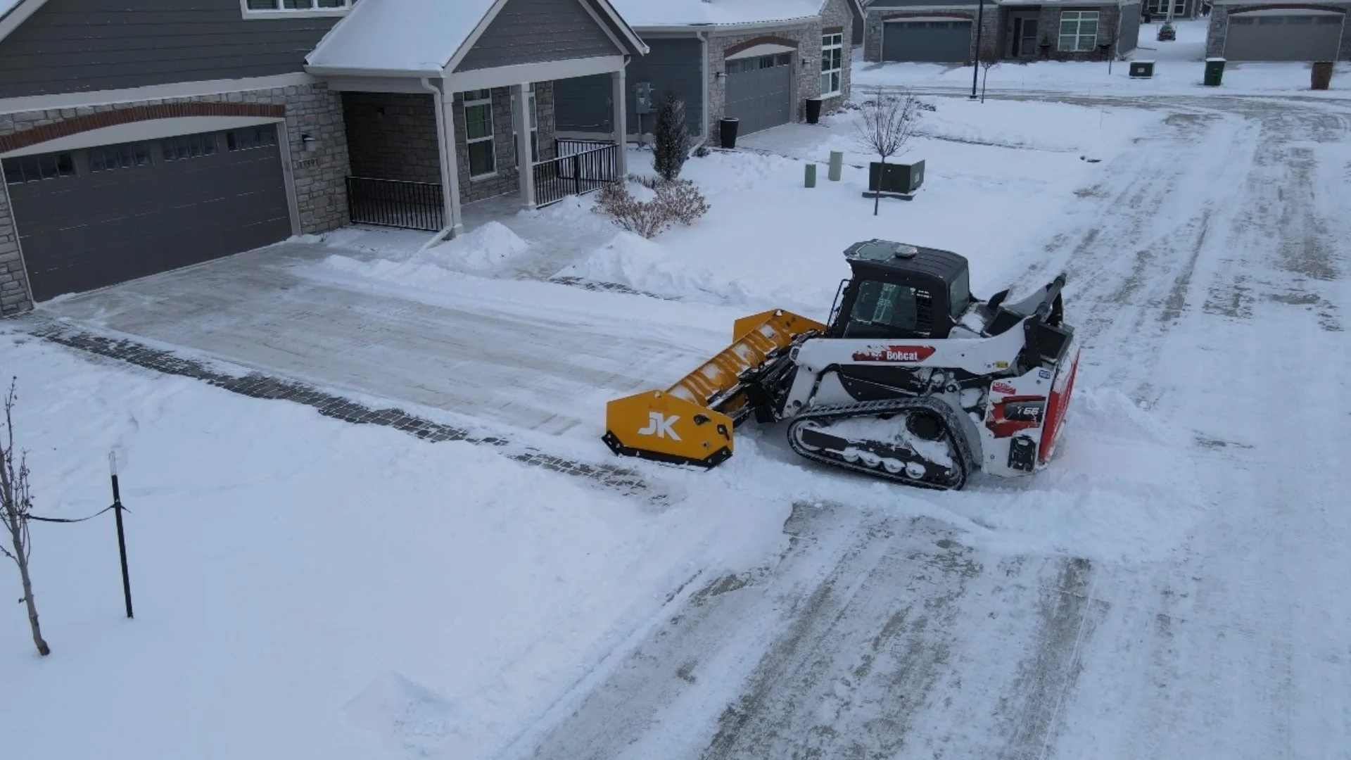 A Bobcat snow plow clearing snow from a residential driveway in a snowy neighborhood, surrounded by houses