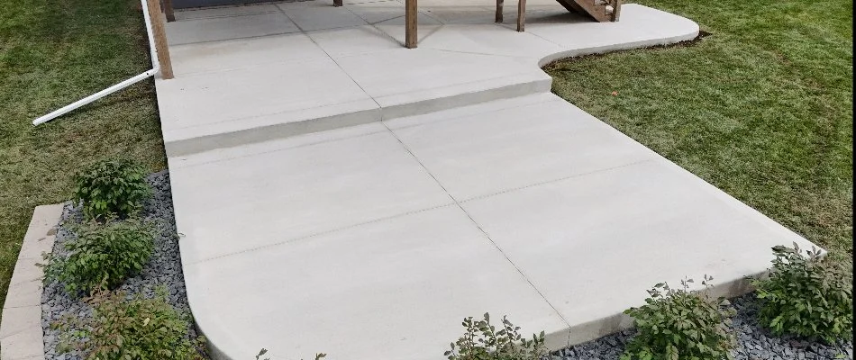 A close-up view of a concrete patio with a staircase leading to a home, featuring a curved edge and landscaped borders with shrubs and decorative stone.