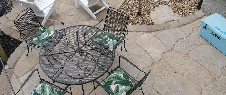 Flagstone patio in Waukee, IA with a fire pit surrounded by white Adirondack chairs, a dining table with tropical cushions, a young tree, and a landscaped backyard featuring a hammock and string lights.