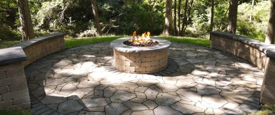 A circular outdoor fire pit surrounded by stone patio seating, set in a scenic forested backyard with lush greenery in Waukee, IA.