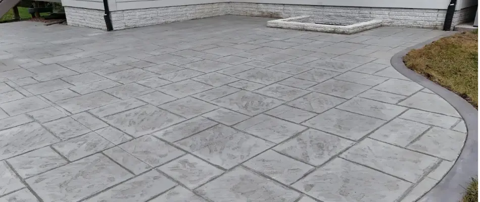 Close-up of a textured concrete patio with a patterned design, showing gray tiles and a smooth edge, adjacent to a grassy area.